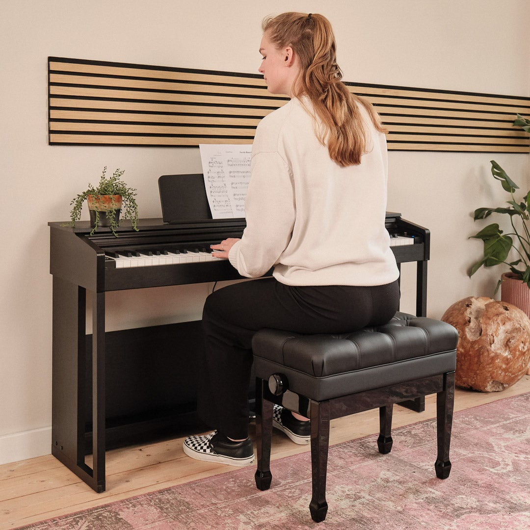 A woman plays a black keyboard piano with sheet music on it, seated on a stool.