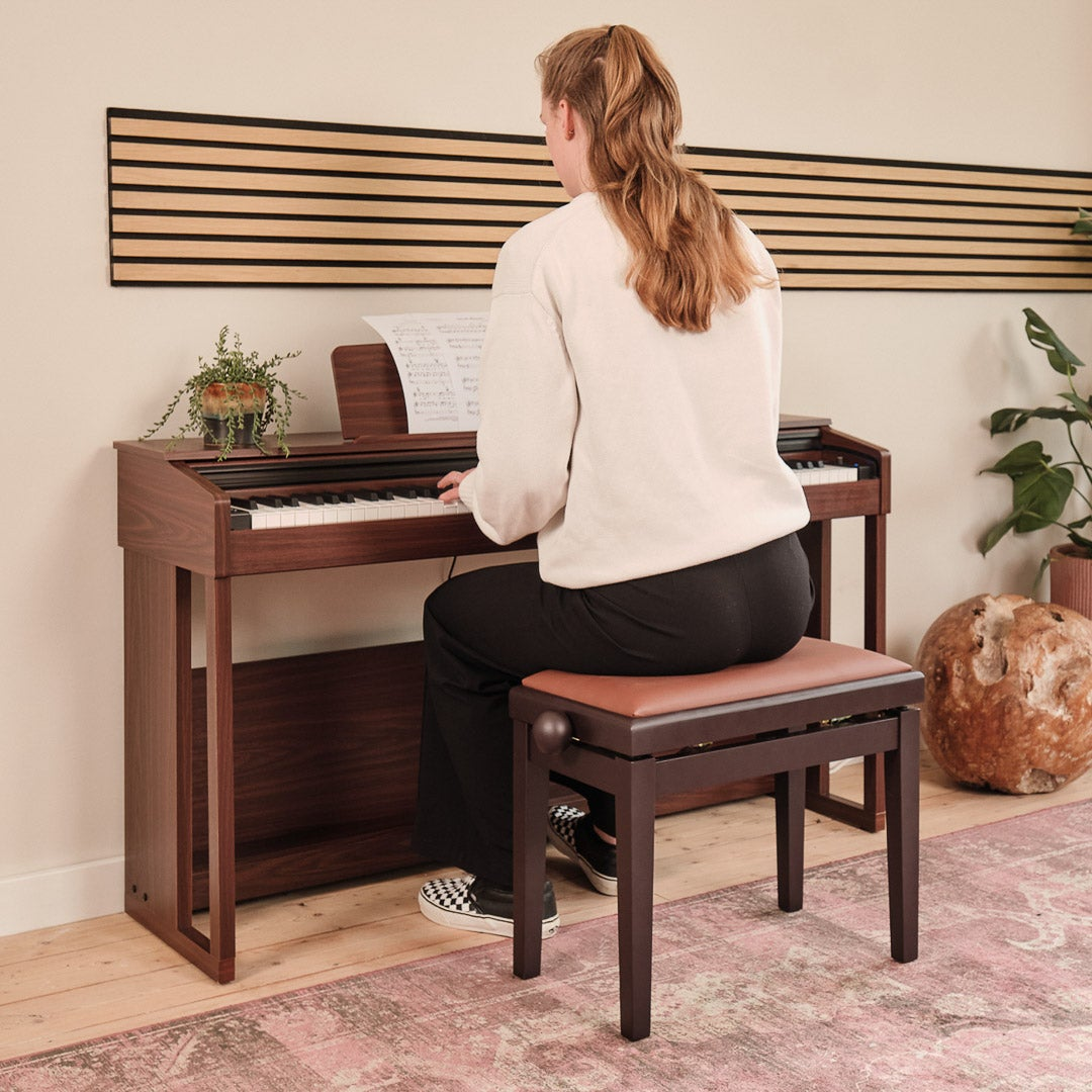 A woman plays the piano, sitting on a brown stool in front of a piano.