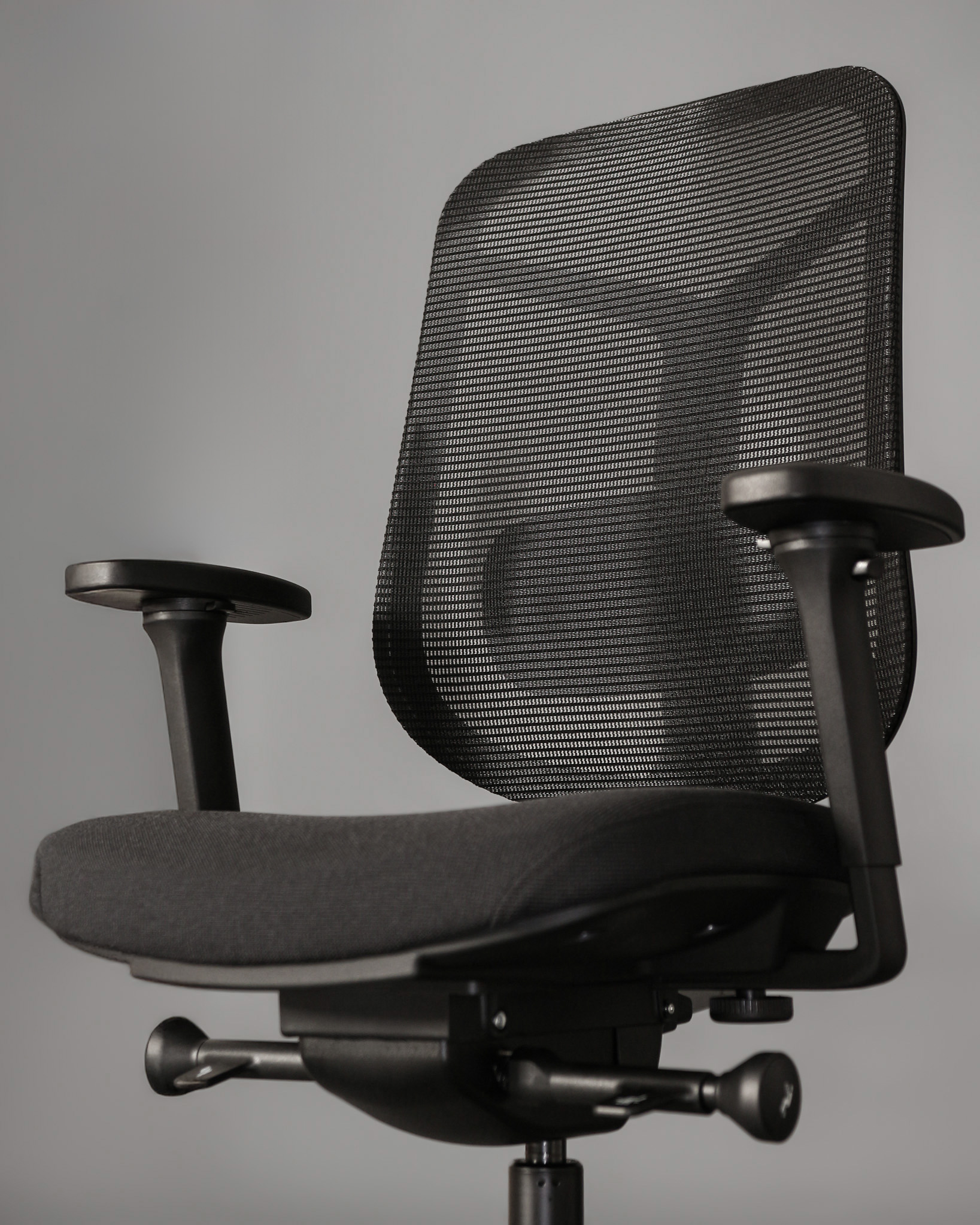 Czarny fotel biurowy z siatkowanym oparciem i podłokietnikami na szarym tle.