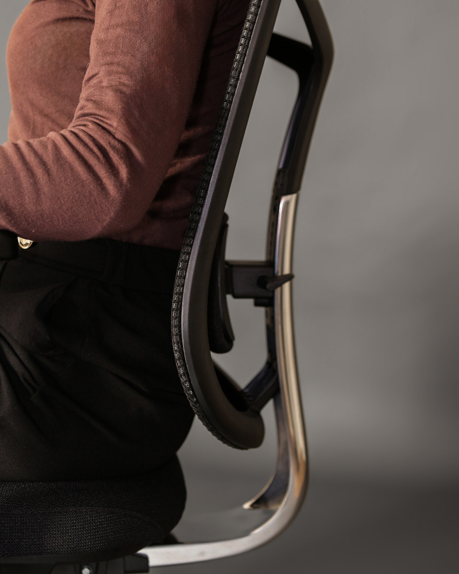 Osoba w brązowym swetrze i czarnych spodniach siedzi na krześle biurowym.