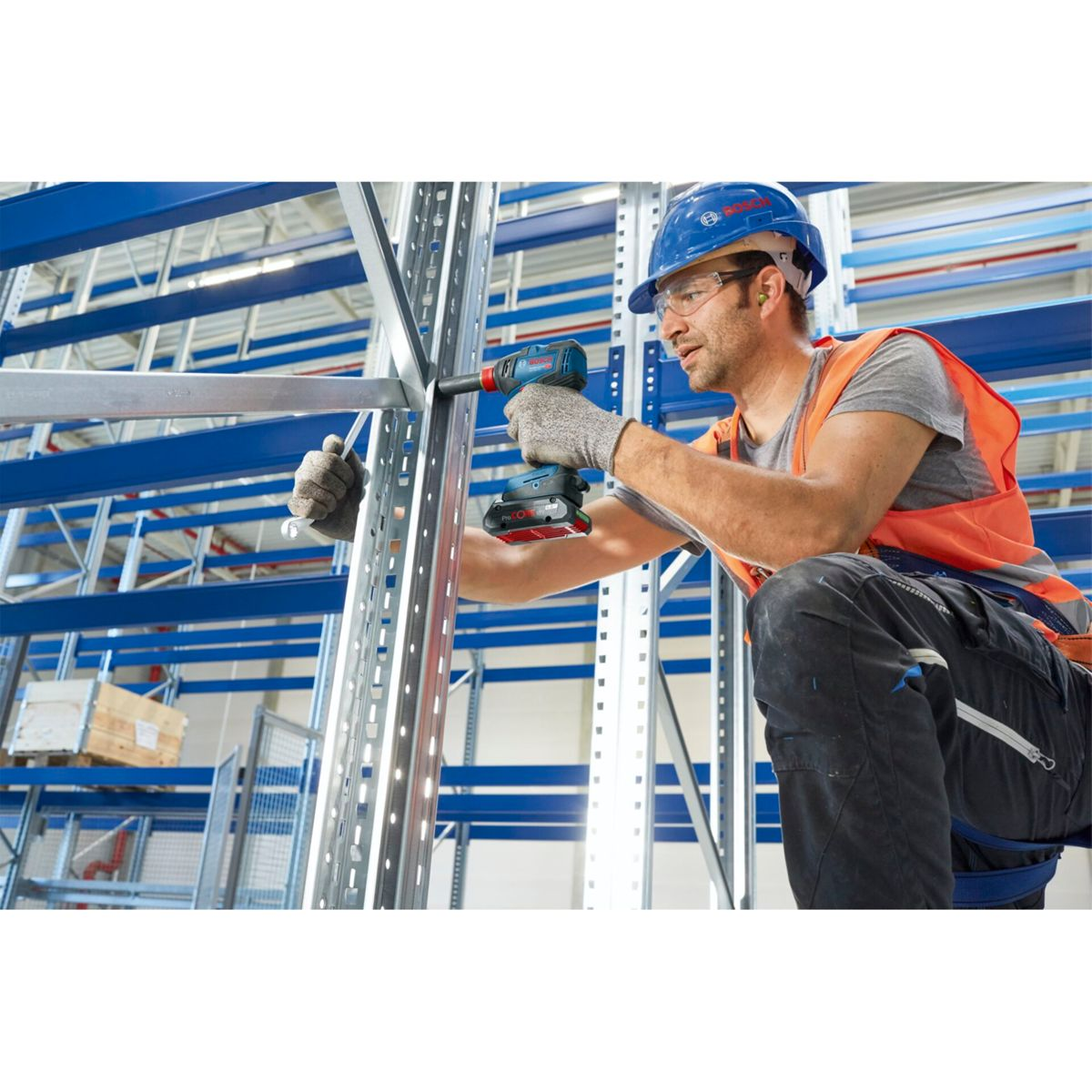 Un uomo con casco e gilet usa un trapano a batteria su uno scaffale metallico.