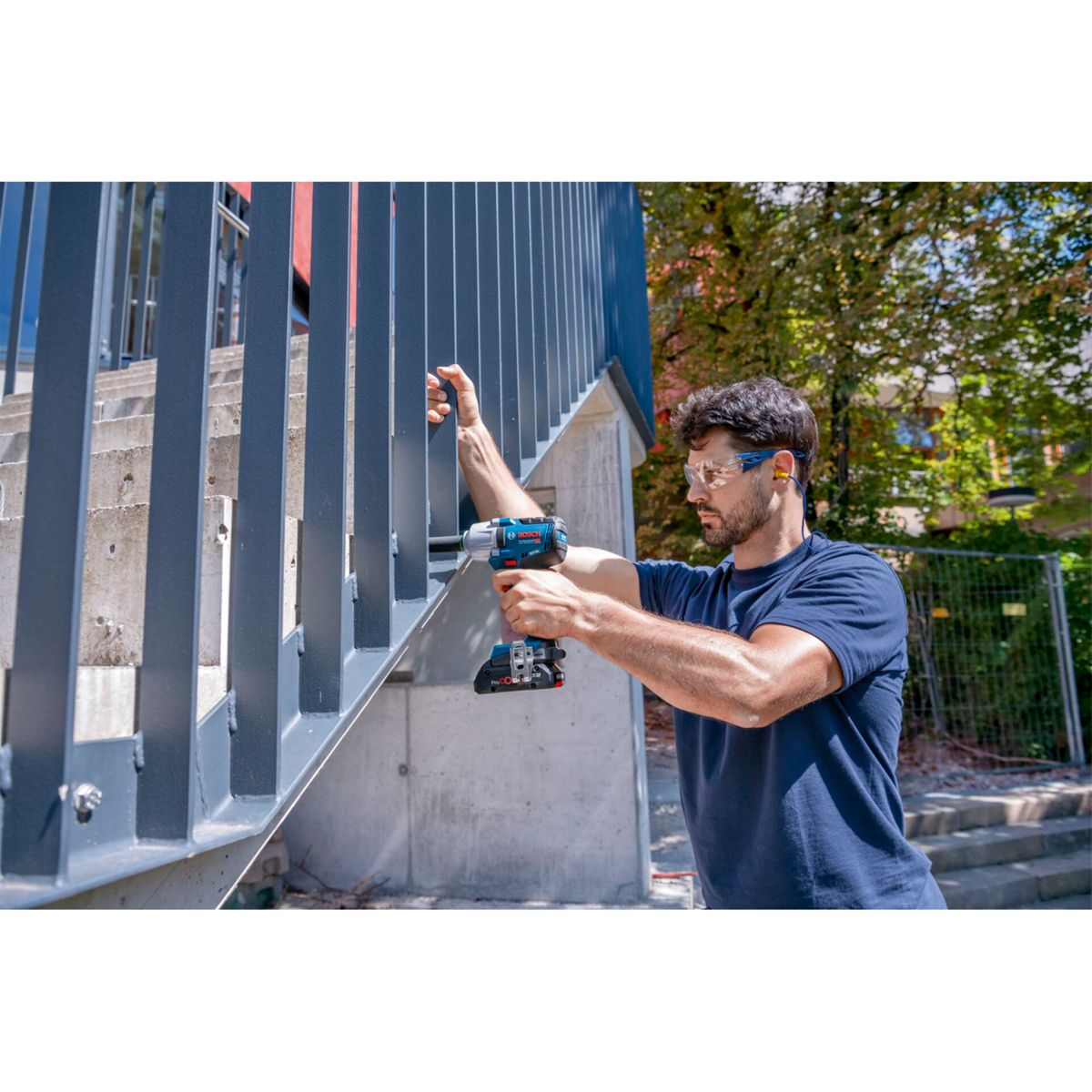 Mężczyzna w okularach ochronnych wierci w metalowych schodach niebieską wiertarko-wkrętarką.