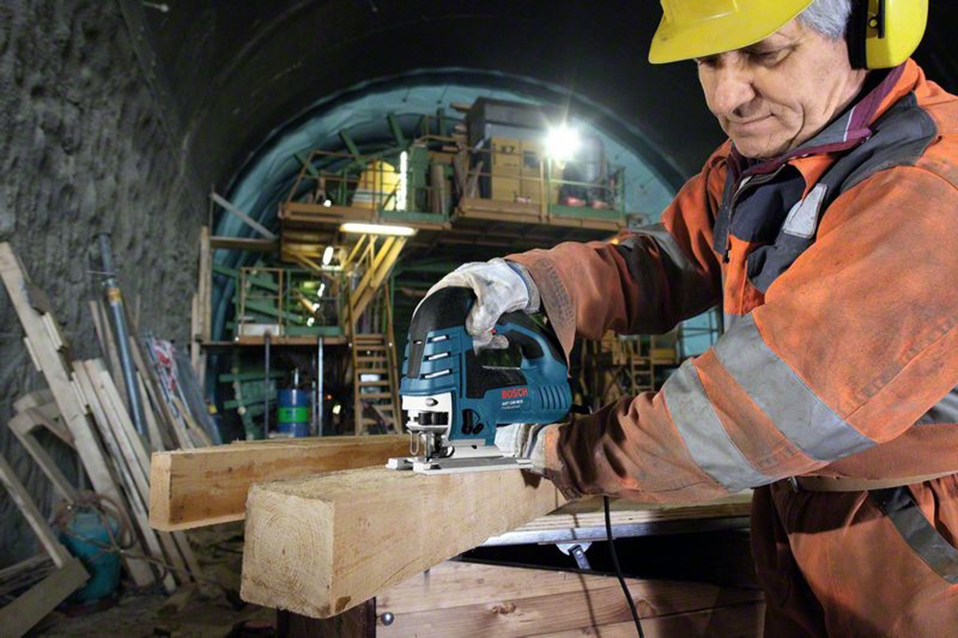 Ein Mann in orangefarbener Arbeitskleidung und gelbem Helm benutzt eine Stichsäge an Holzbalken in einem Tunnel.