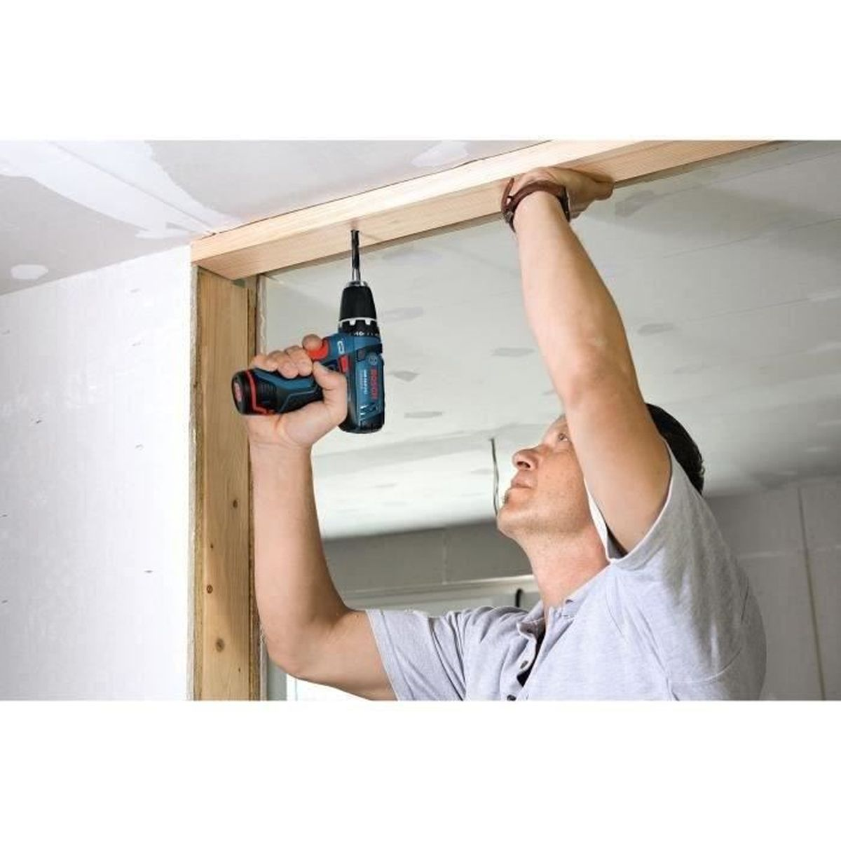 Mężczyzna używa niebieskiej wiertarki Bosch do wkręcania drewnianej belki w sufit.