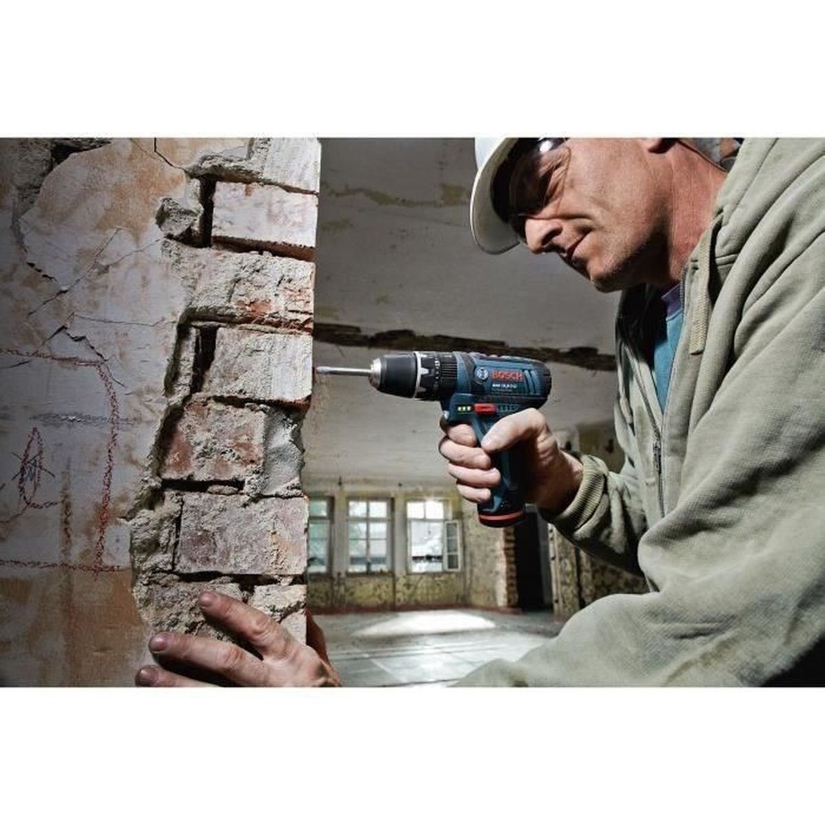 Mężczyzna z wiertarką wierci w ścianę z cegły, plac budowy w tle.
