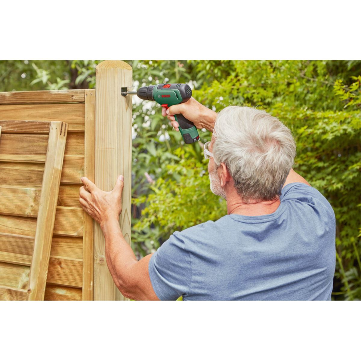 Mężczyzna przykręca drewniany słup do ogrodzenia wkrętarką.