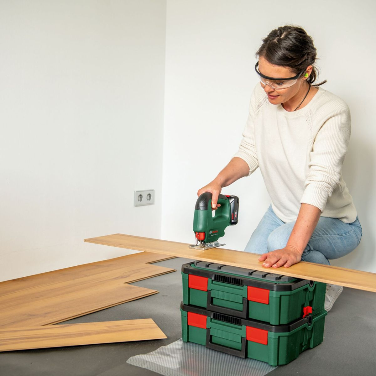 Eine Frau mit Schutzbrille sägt Holzplanken mit einem grünen Stichsäge.