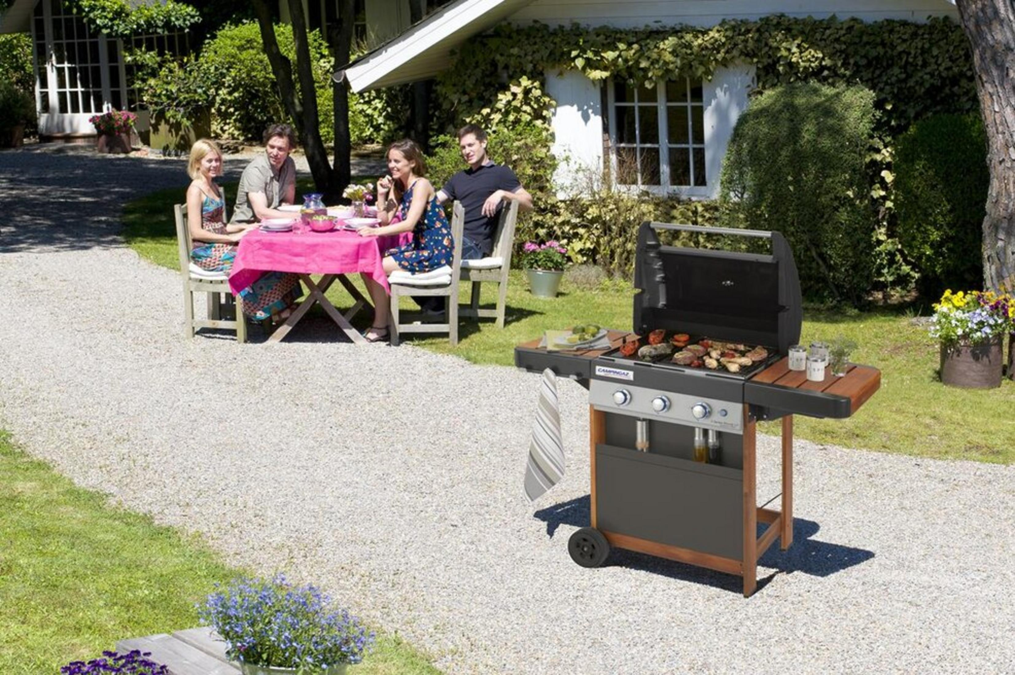 Vier Personen sitzen an einem rosafarbenen Tisch im Freien, während ein Grill in der Nähe steht.