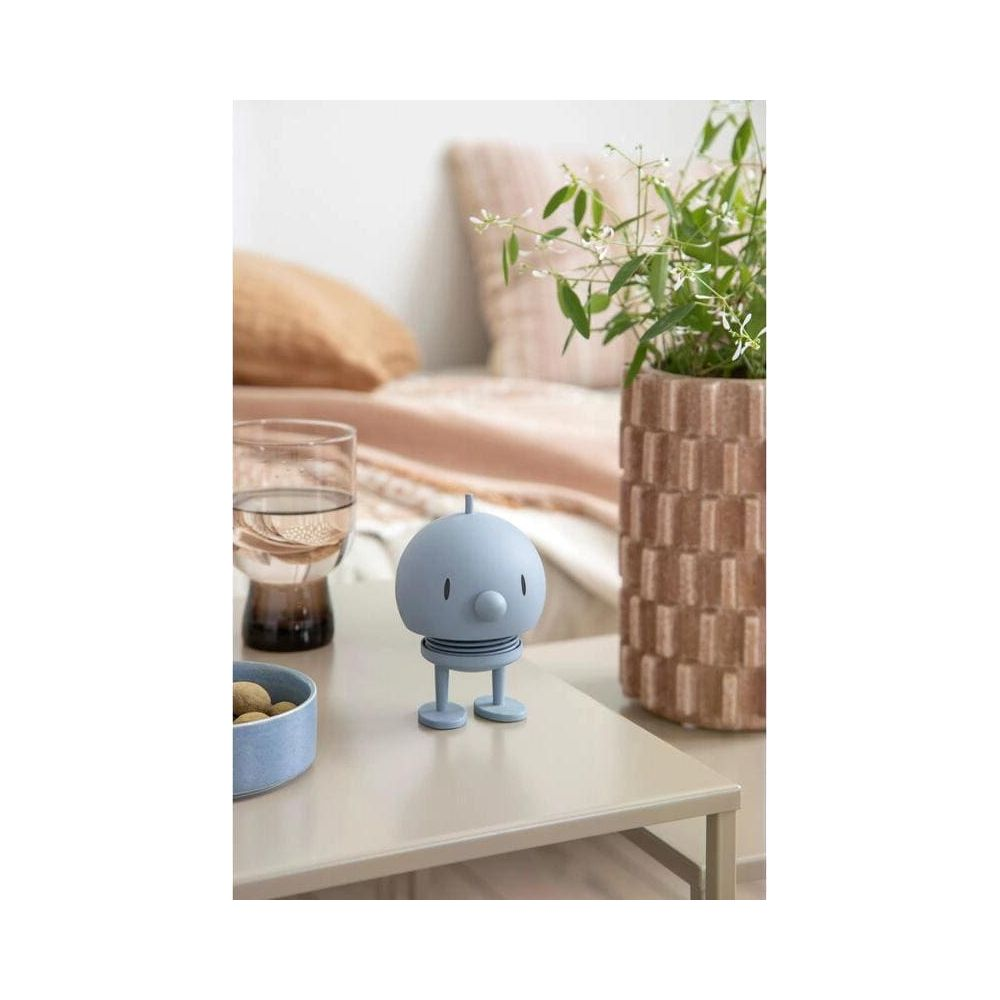 Ein blauer Roboter steht auf einem Tisch neben einem Glas Wasser und einer Vase mit Pflanzen.