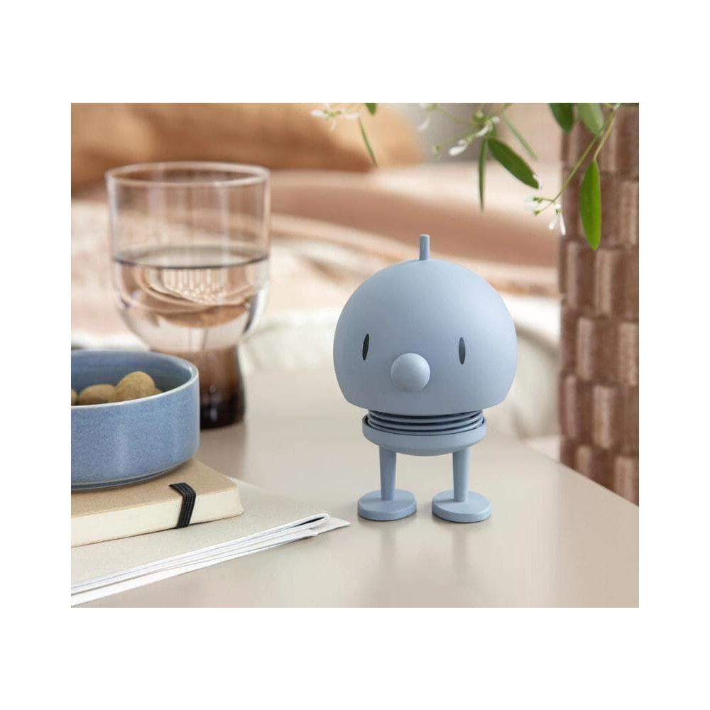 Eine hellblaue Roboterfigur steht auf einem Tisch neben einem Glas Wasser und einer Schale.