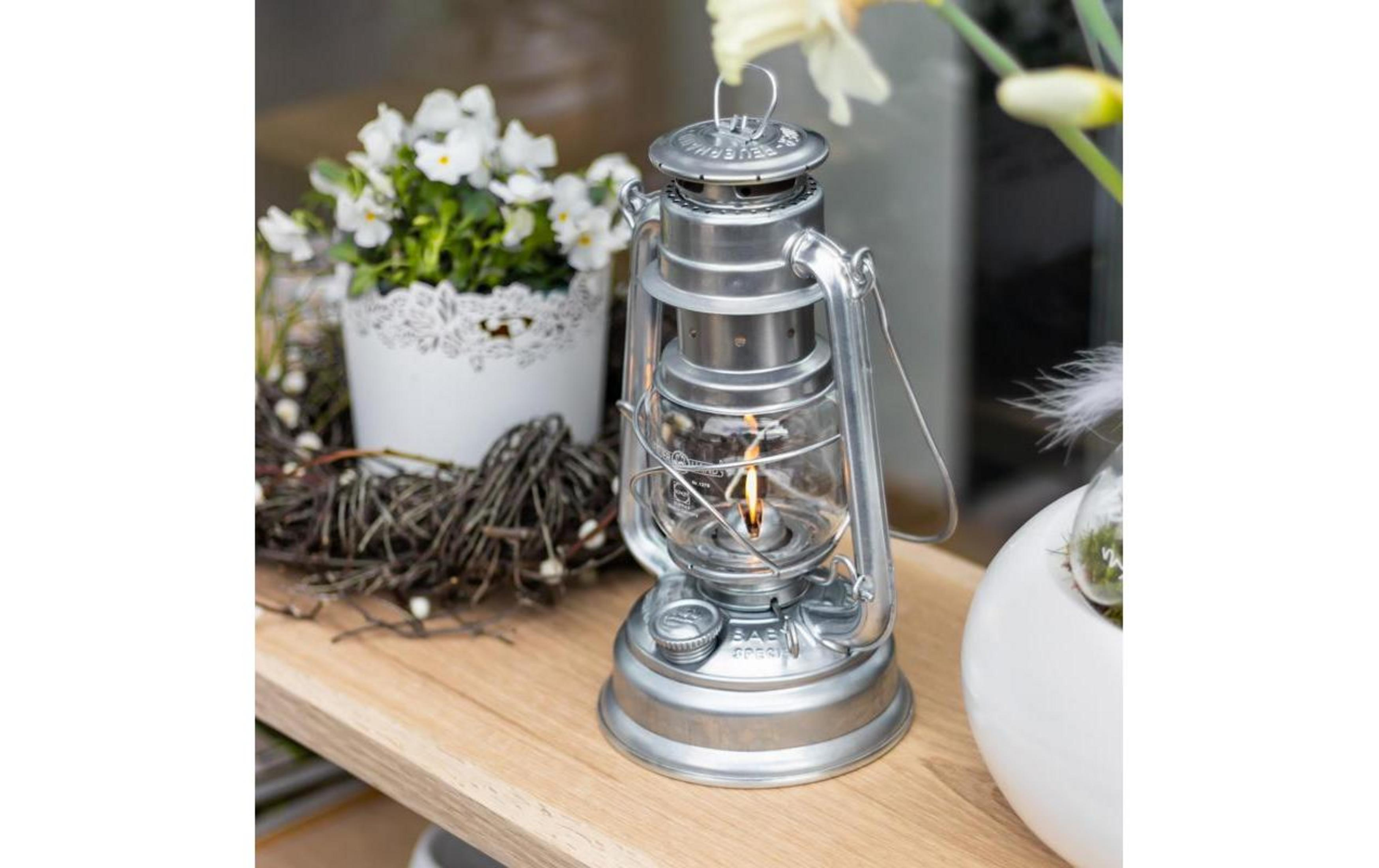 Eine silberne Petroleumlampe brennt auf einem Holzregal neben weißen Blumen.