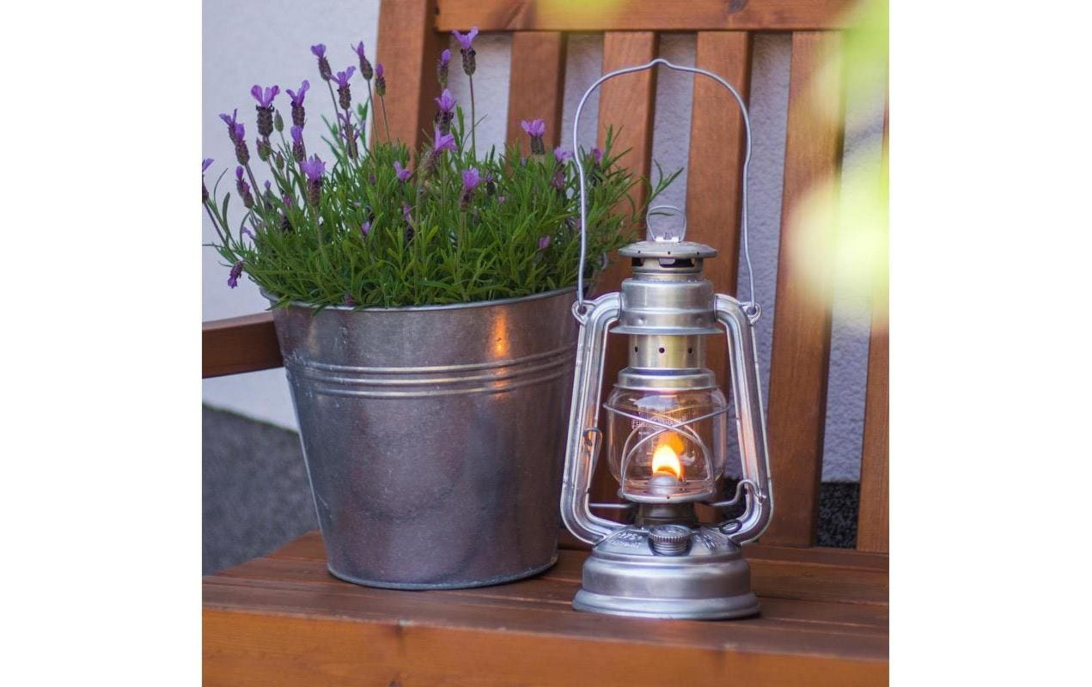 Lavendel in Zinktopf neben brennender Petroleumlampe auf Holzbank.