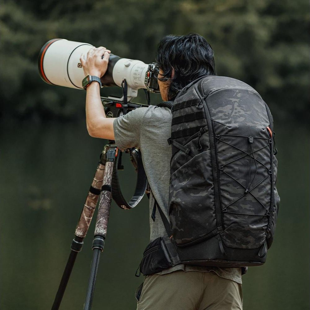 Un photographe avec un sac à dos utilise un grand appareil photo sur trépied, en extérieur.