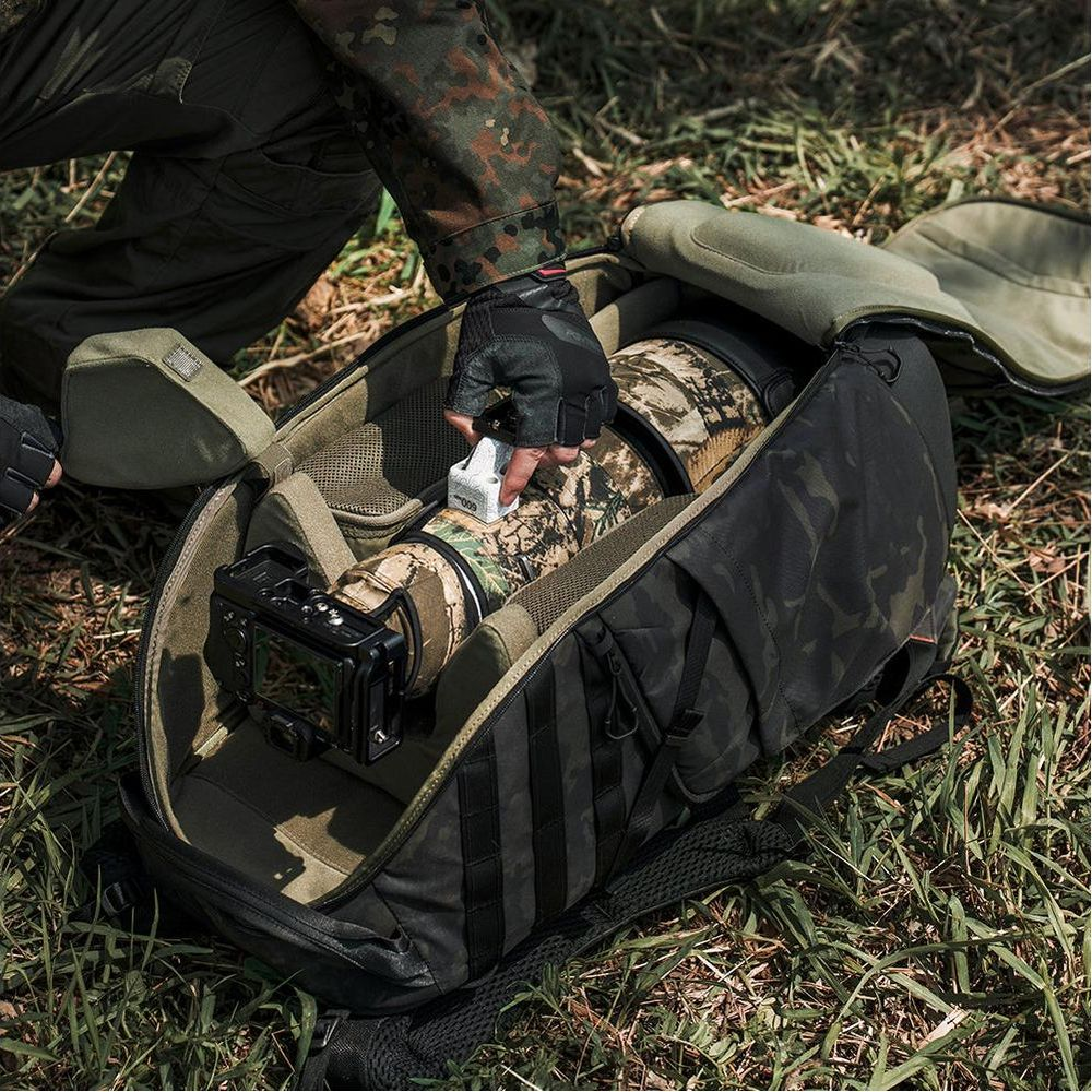Une main gantée place un objet blanc sur un appareil photo avec objectif camouflage dans un sac à dos.