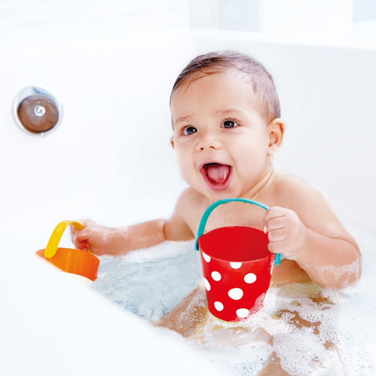 Un bébé souriant est assis dans une baignoire remplie d'eau et de bulles, tenant un seau rouge à pois.
