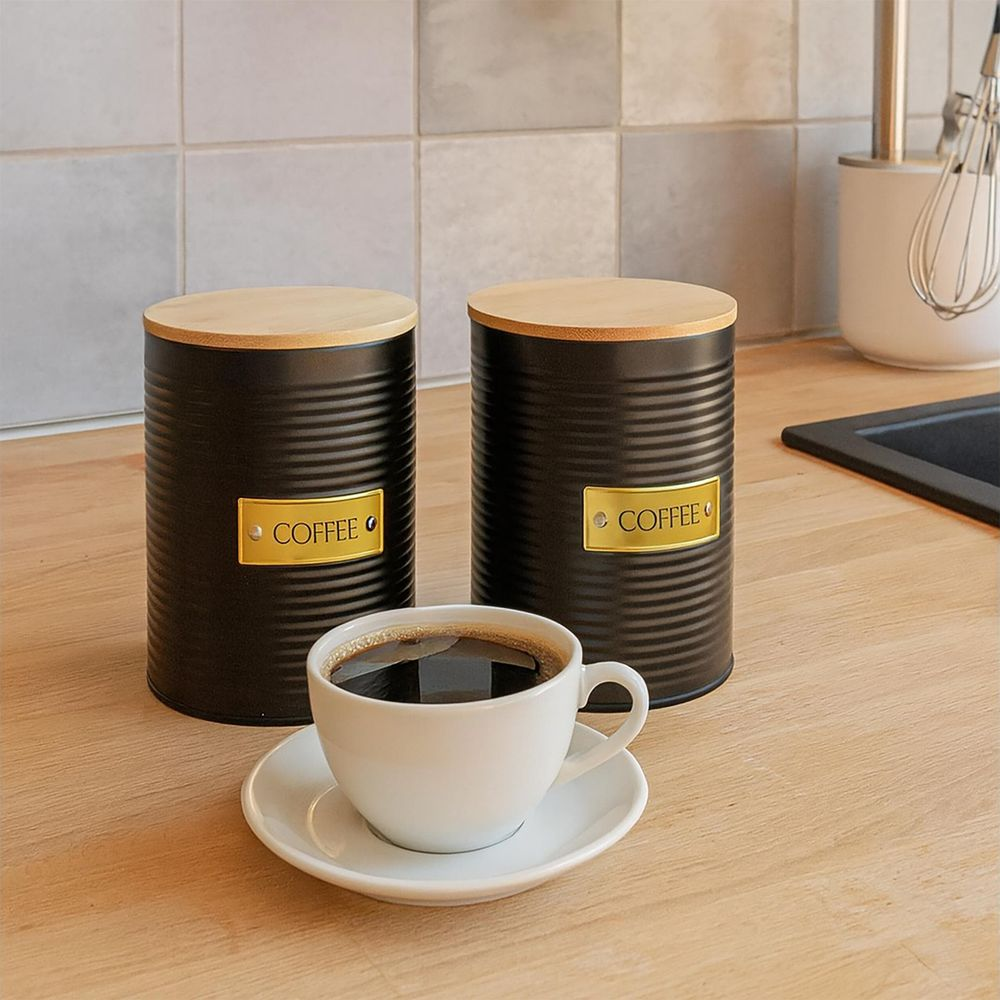 Zwei schwarze Kaffeedosen mit Holzdeckeln und eine weiße Kaffeetasse auf einem Holztresen.