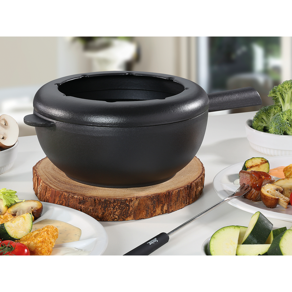 Schwarzer Fondue-Topf auf Holzscheibe, mit Fleisch, Gemüse und Dips.