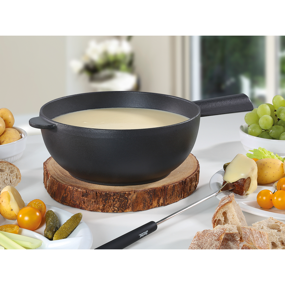Ein schwarzer Fonduetopf mit cremiger Soße, daneben Brot, Trauben, Kartoffeln und Gurken.