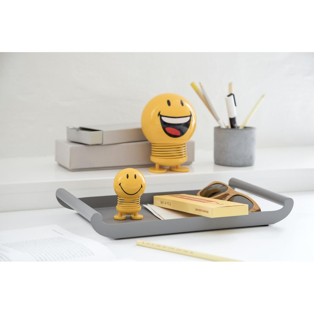 Figure sorridenti gialle e occhiali da sole su un vassoio grigio, con cancelleria sullo sfondo.