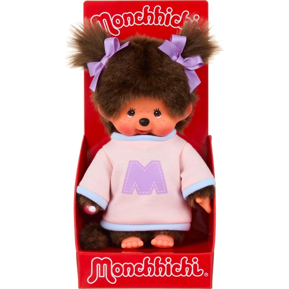 Una bambola Monchhichi con pelo marrone, fiocchi viola e maglietta rosa con 'M' nella scatola.
