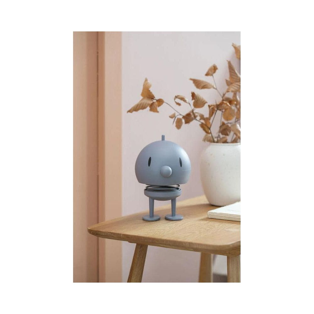 Graue Figur mit Feder, auf einem Holztisch neben einer Vase mit Zweigen.