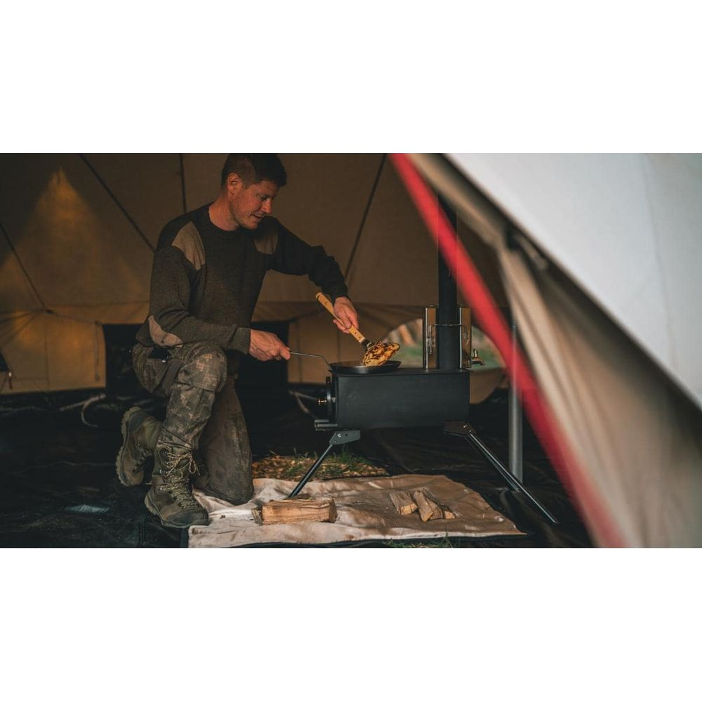 Un uomo si inginocchia in una tenda, cucinando su una stufa a legna portatile.