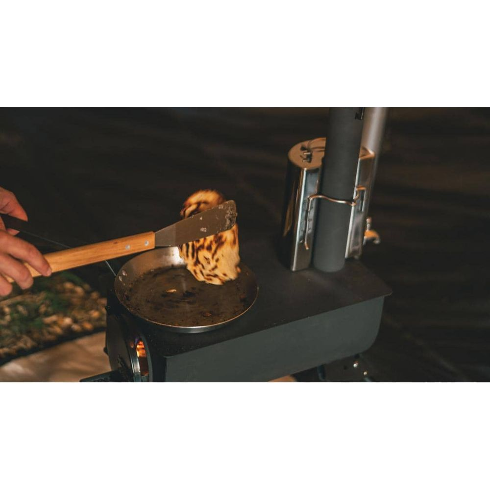 Una mano usa una pinza per girare un pancake dorato in una piccola padella su un fornello da campeggio nero.