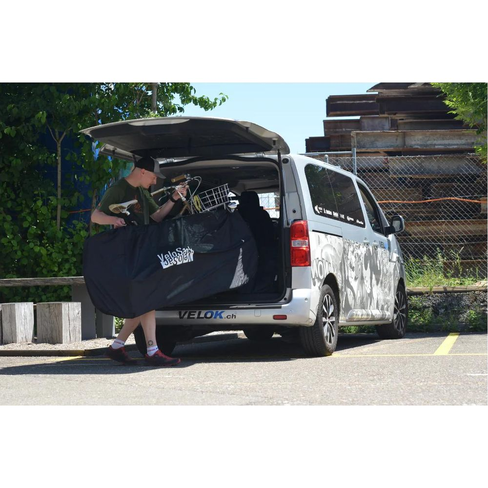 Un homme emballe un vélo dans un sac noir à côté d'une camionnette argentée avec autocollants.