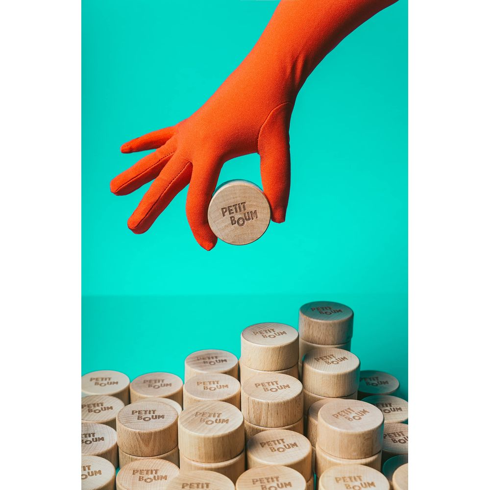 Eine orangefarbene Hand hält einen Holzzylinder mit der Aufschrift „PETIT BOUM“.