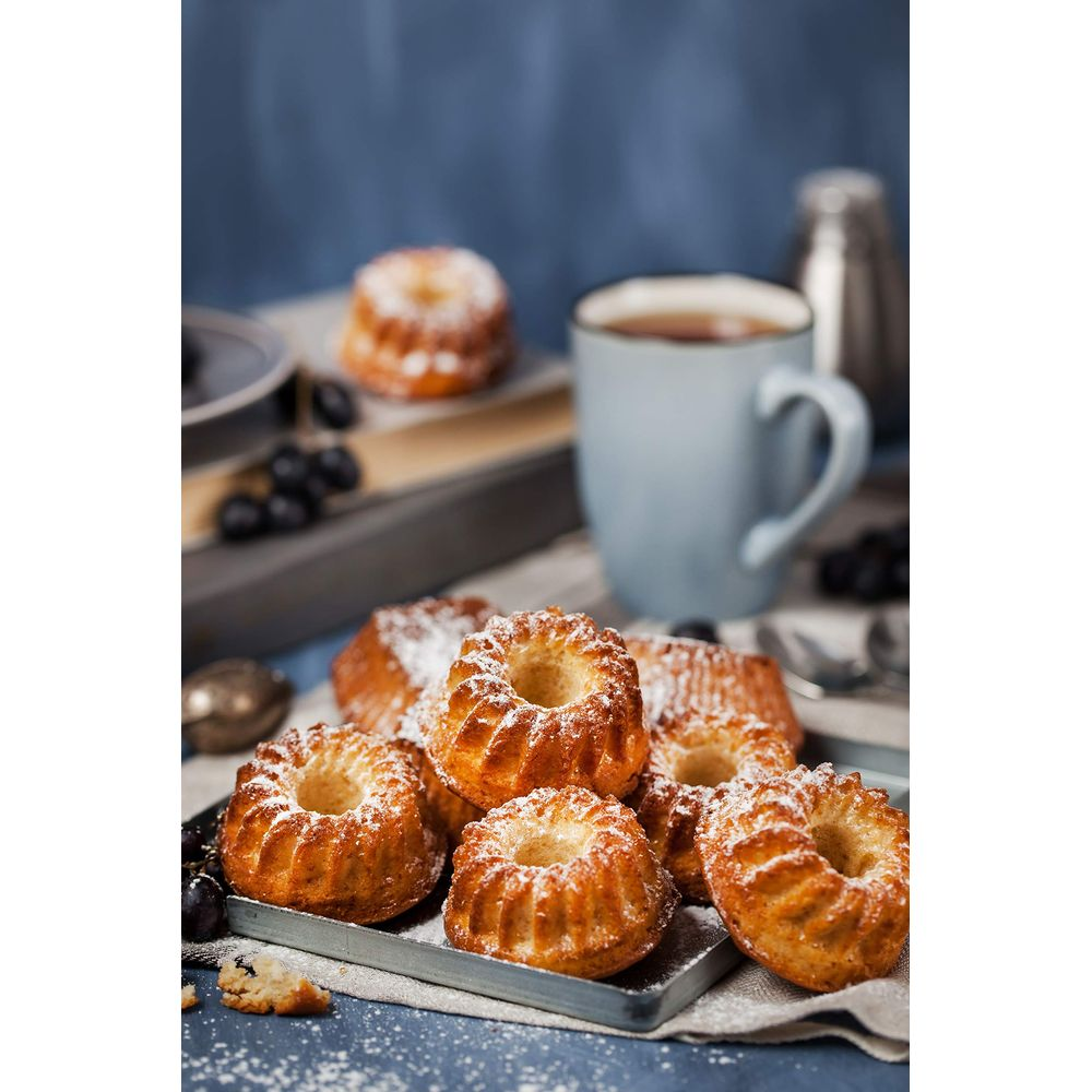 Małe babeczki typu bundt posypane cukrem pudrem i jagodami na tacy obok filiżanki herbaty.