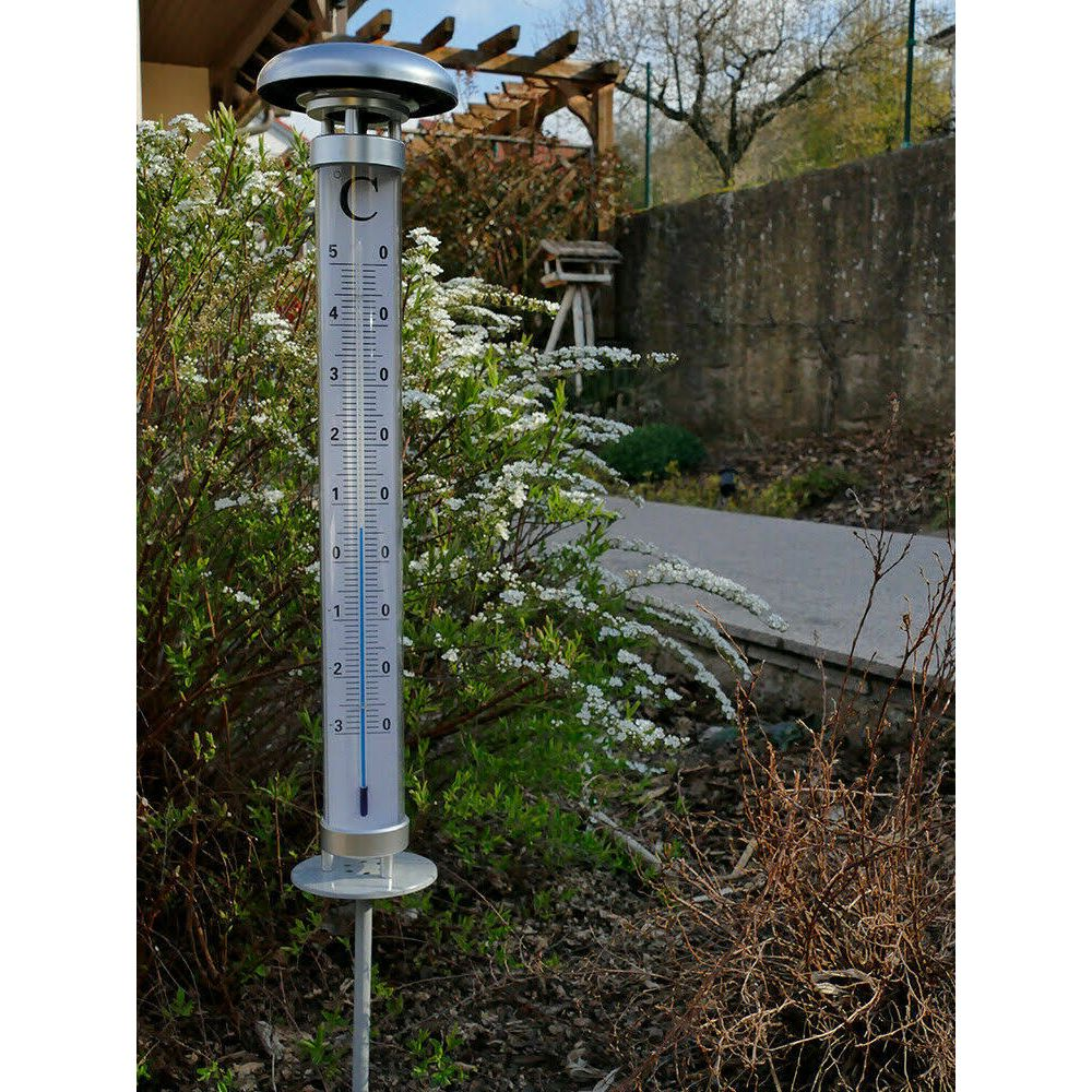 Ein Thermometer steht im Garten neben blühenden Sträuchern.
