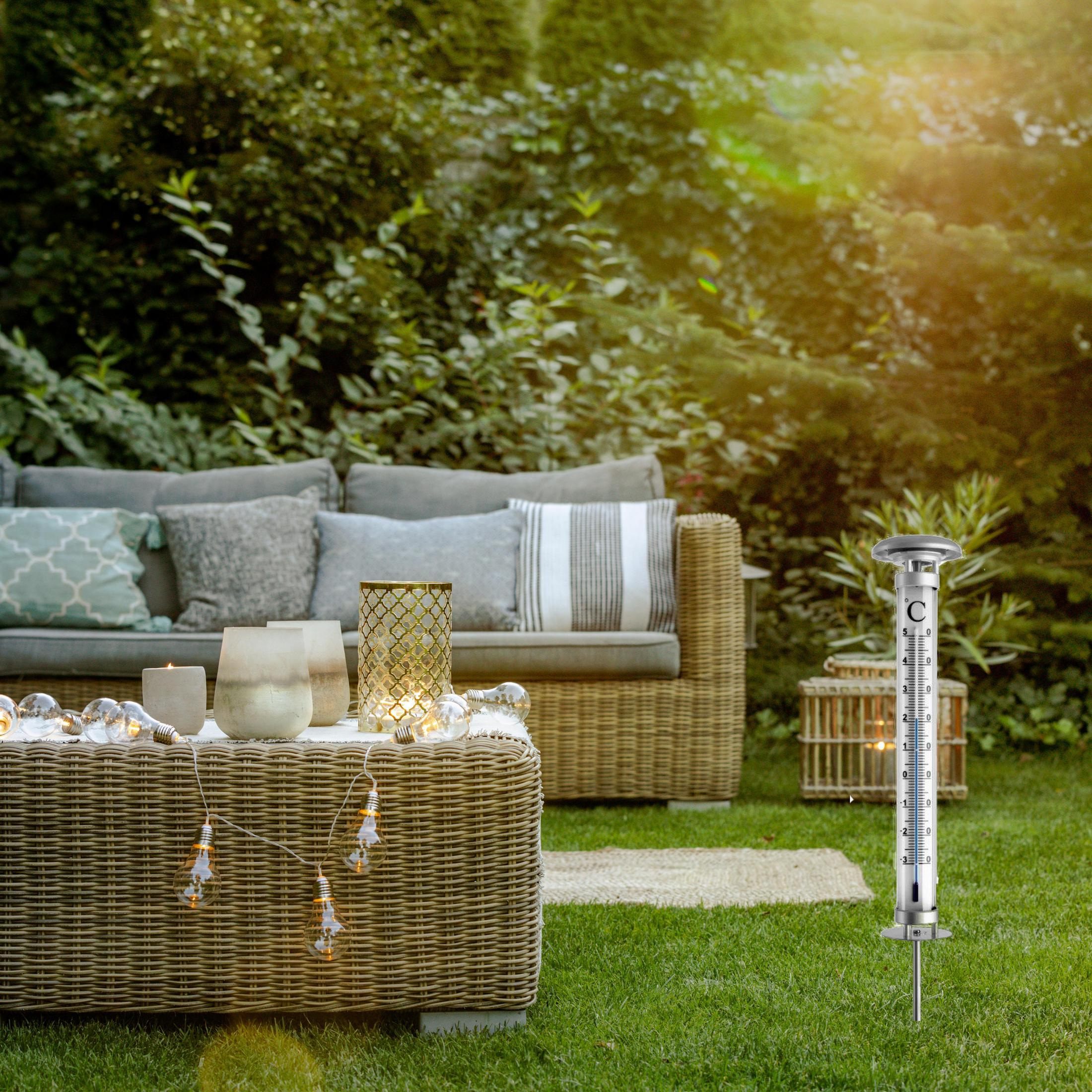 Ein Thermometer steht im Gras neben einem Gartensofa und einem Tisch mit Lichtern.
