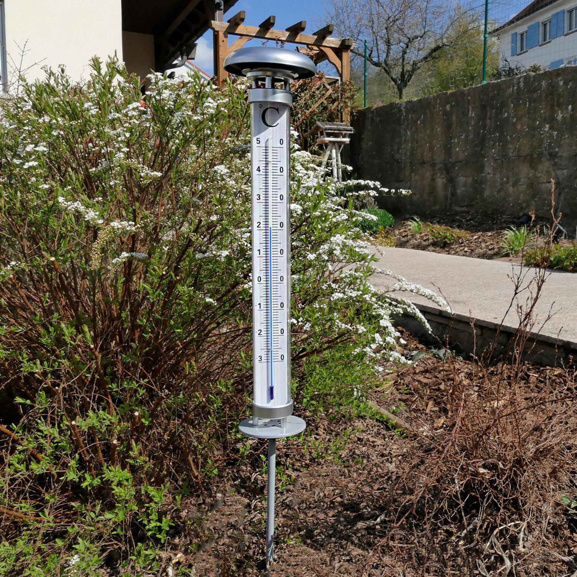 Ein Thermometer steht im Garten neben blühenden Büschen.