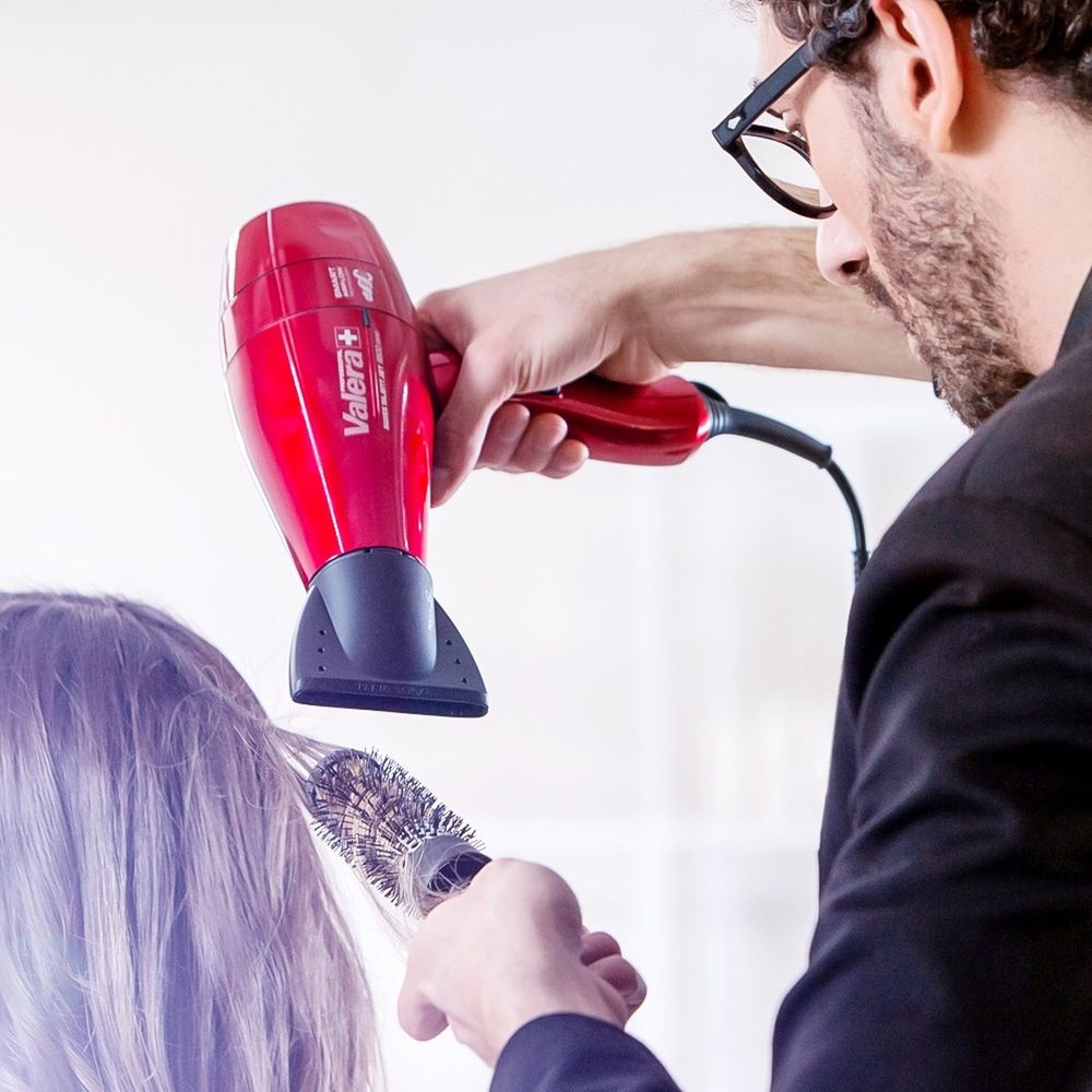 Ein Friseur trocknet das Haar einer Kundin mit einem roten Föhn und einer Rundbürste.