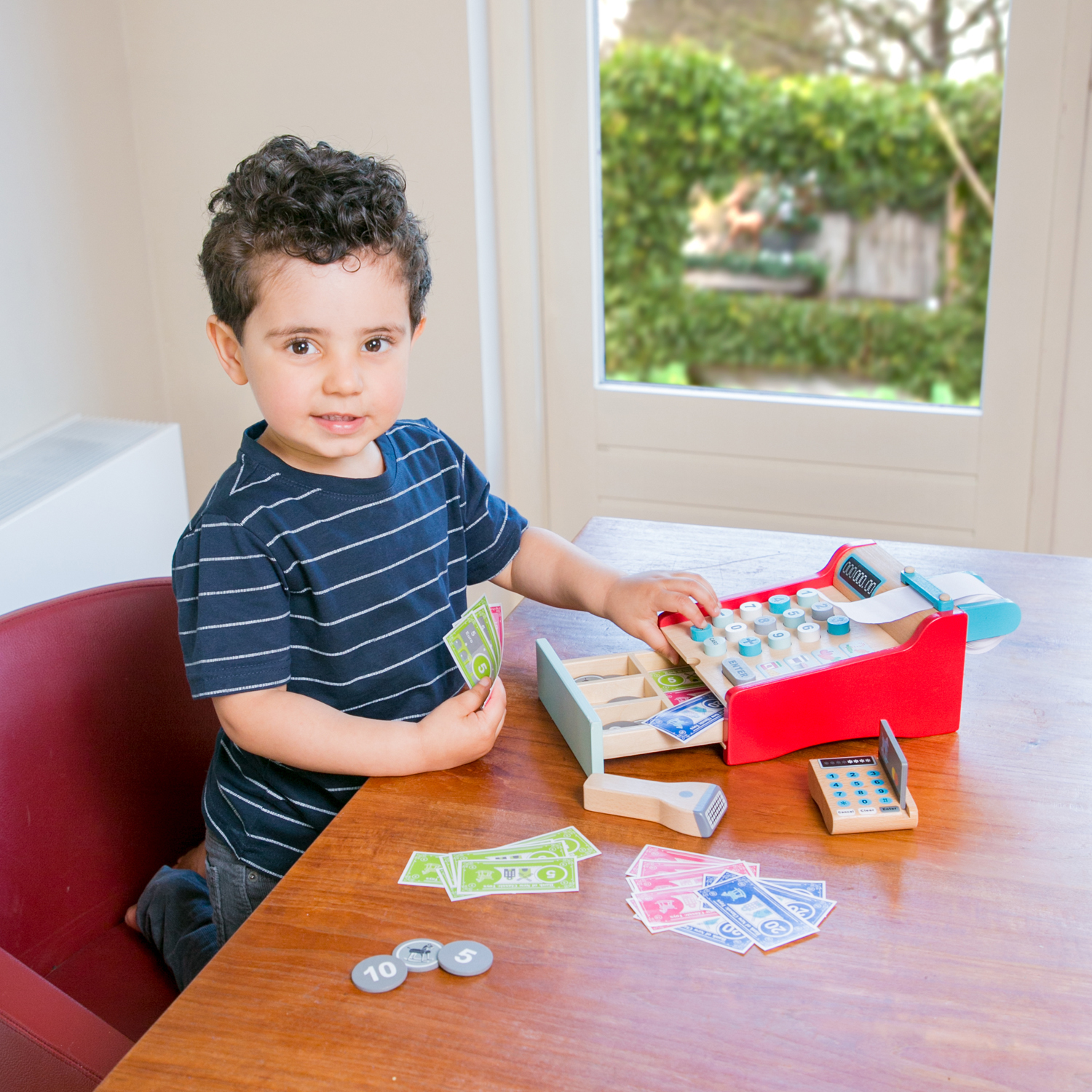 Ein kleiner Junge spielt mit einer Spielzeugkasse und Spielgeld an einem Holztisch.
