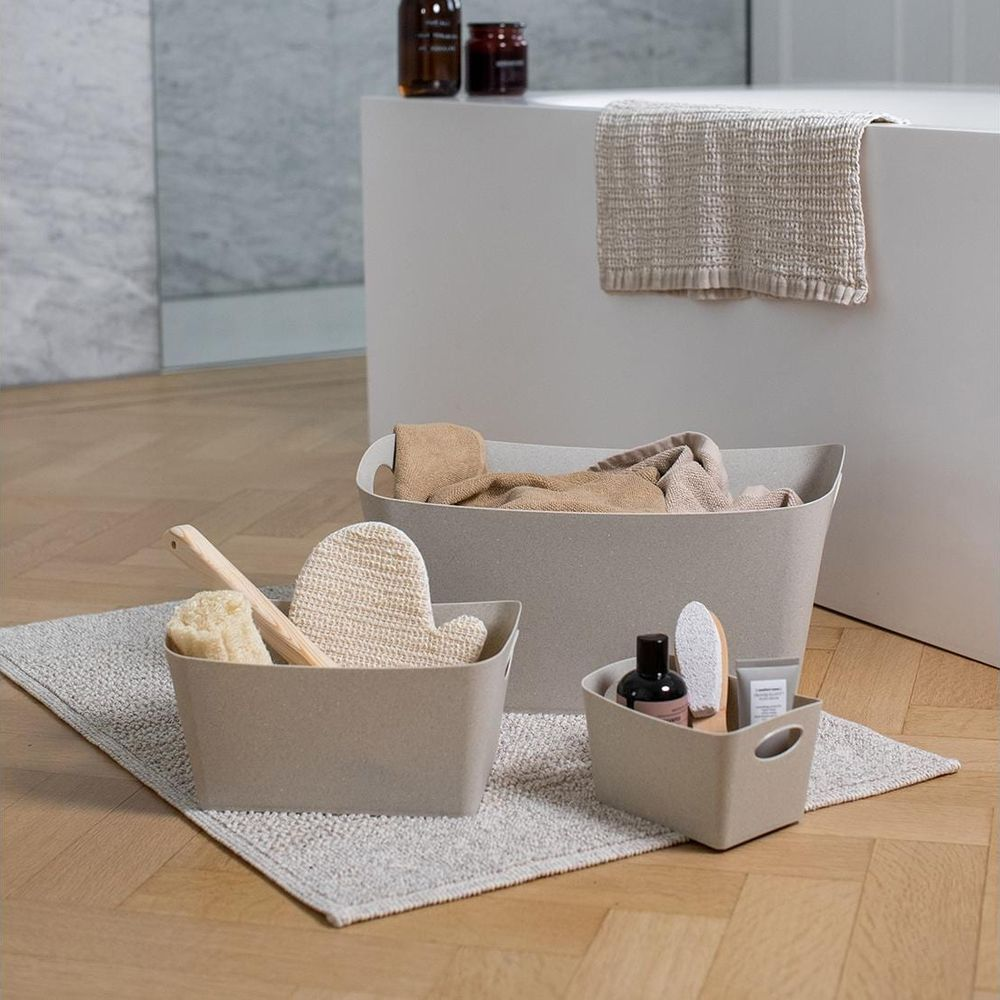 Beige Körbe mit Badezimmerartikeln auf Holzboden, Handtuch im Hintergrund.