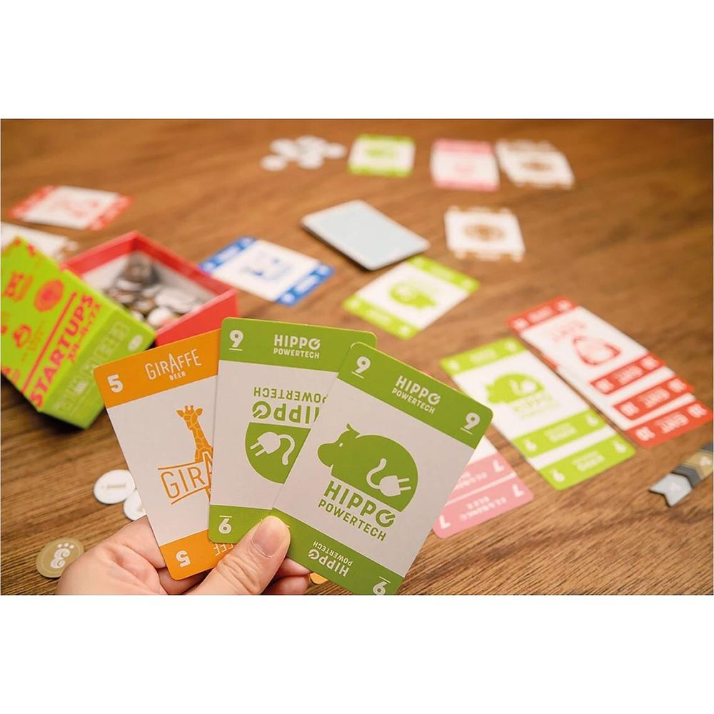 Eine Hand hält Spielkarten mit Giraffen- und Nilpferdmotiven. Andere Karten und eine Schachtel liegen auf einem Holztisch.
