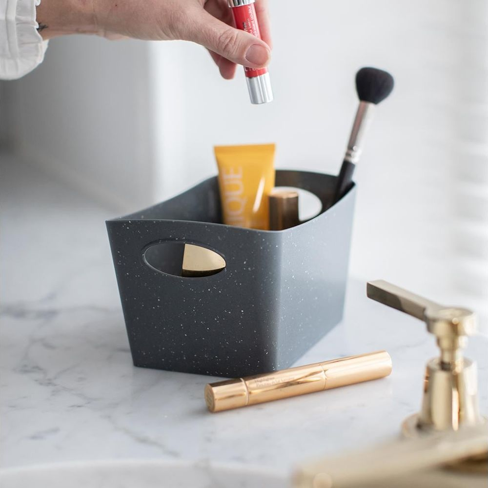Eine Hand legt einen roten Lippenbalsam in einen gesprenkelten grauen Behälter mit Make-up.