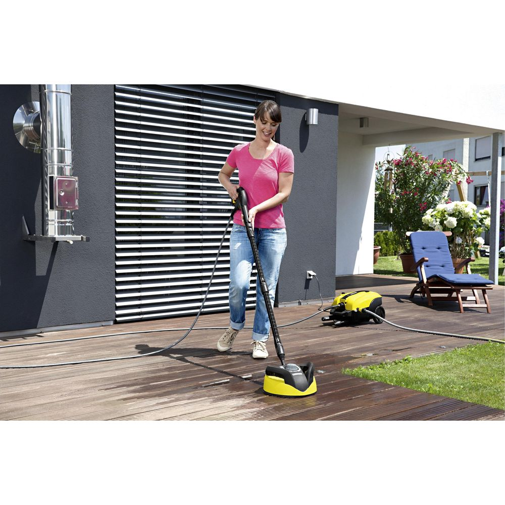 Donna pulisce terrazzo in legno con idropulitrice, macchina gialla sullo sfondo.
