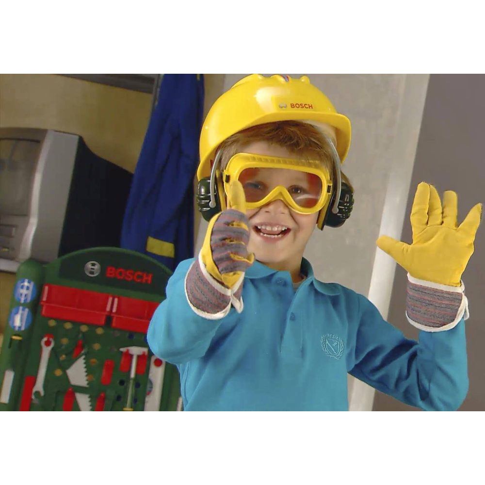 Un bambino indossa casco, occhiali e guanti gialli davanti a una parete degli attrezzi.