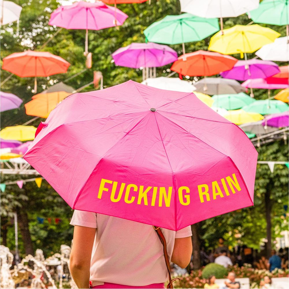 Eine Person hält einen pinken Regenschirm mit gelbem Text. Viele bunte Schirme hängen darüber.