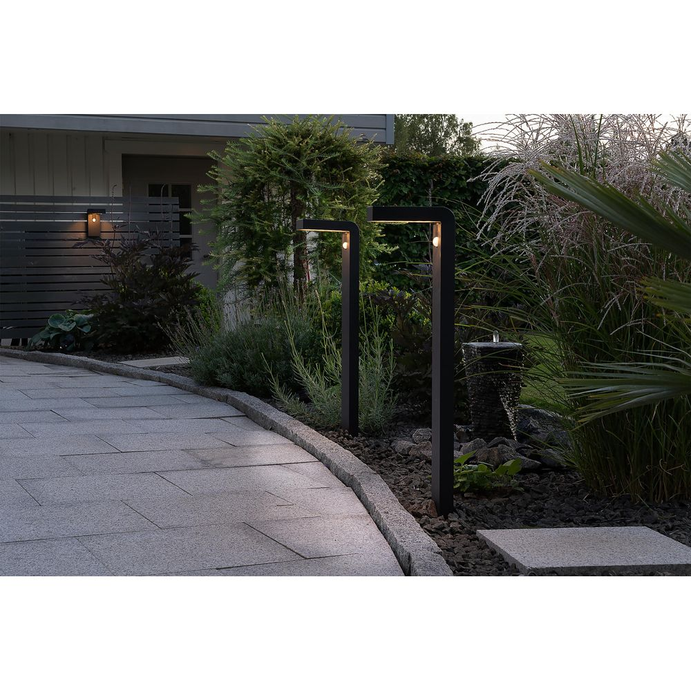 Moderner Gartenweg mit zwei schwarzen Pollerleuchten, kleinem Brunnen und üppigem Grün.
