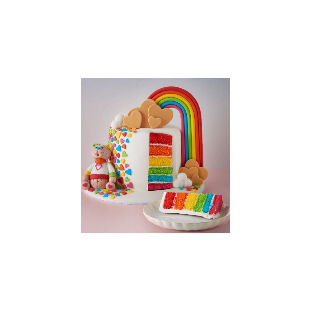 Ein Teddybär neben einem Regenbogenkuchen mit bunten Schichten und Herzdekorationen.