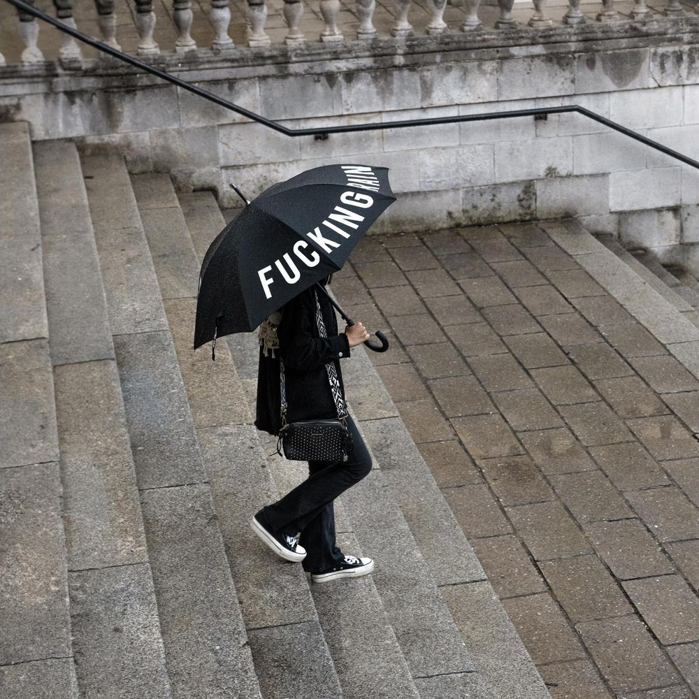 Eine Person mit einem schwarzen Regenschirm mit der Aufschrift „FUCKING RAIN“ geht eine Steintreppe hinunter.