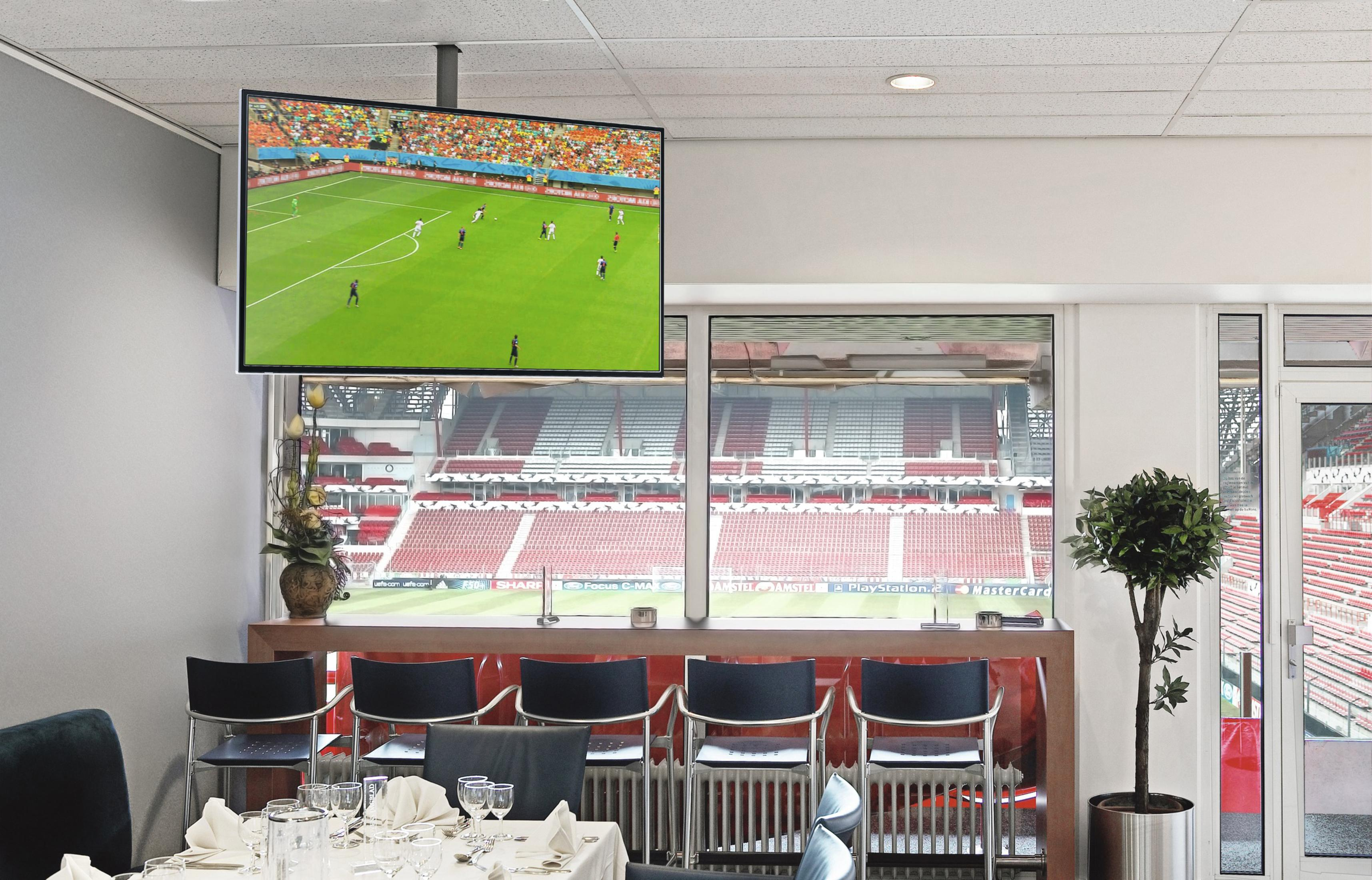 Ein Fernseher zeigt ein Fußballspiel, darunter ein Fenster mit Blick auf ein Stadion.