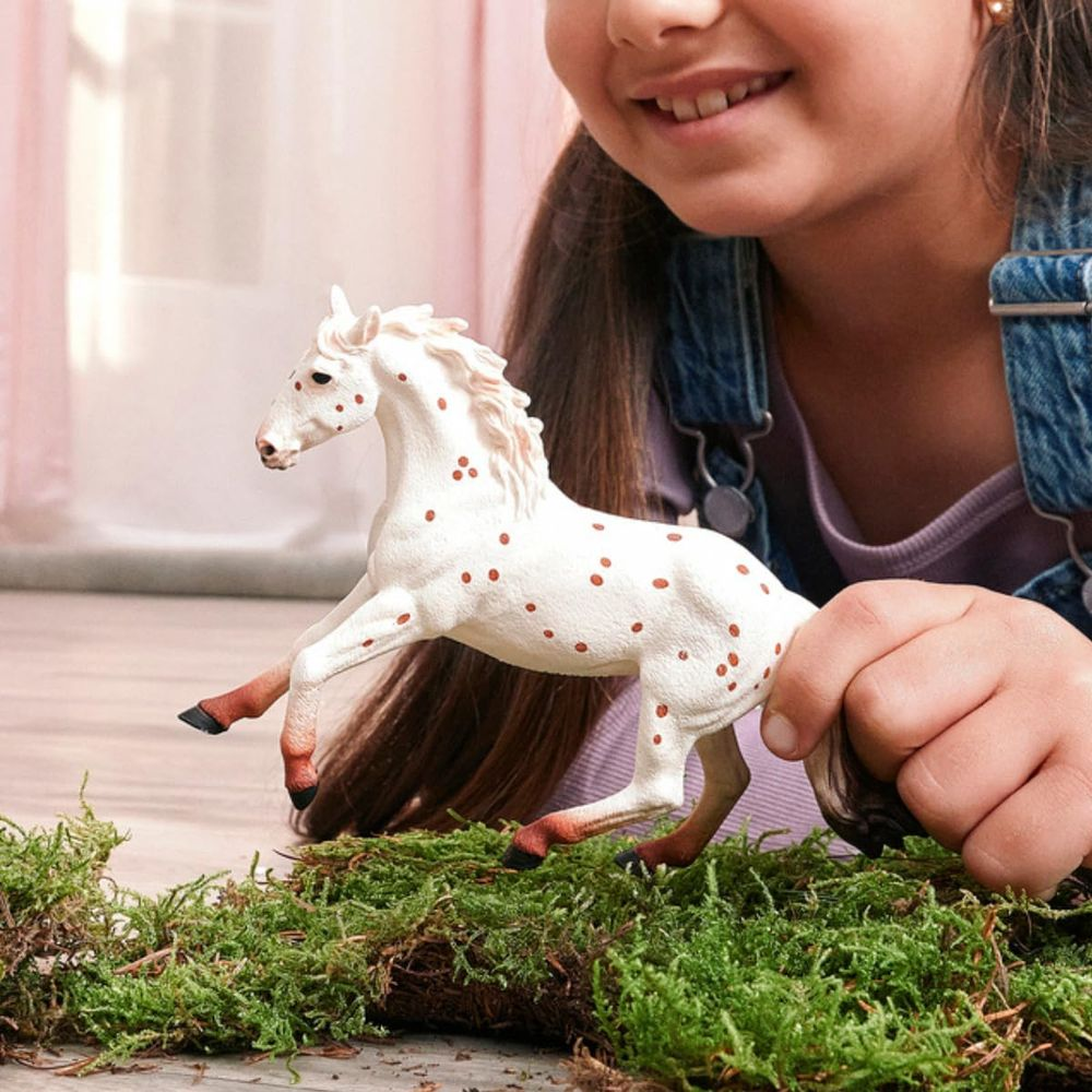 Ein Mädchen hält ein weißes Spielzeugpferd mit braunen Flecken über Moos.