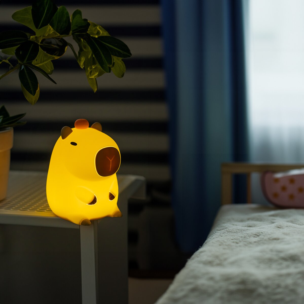 Eine gelbe Capybara-Nachtlampe sitzt auf einem Tisch neben Pflanzen und einem Bett.