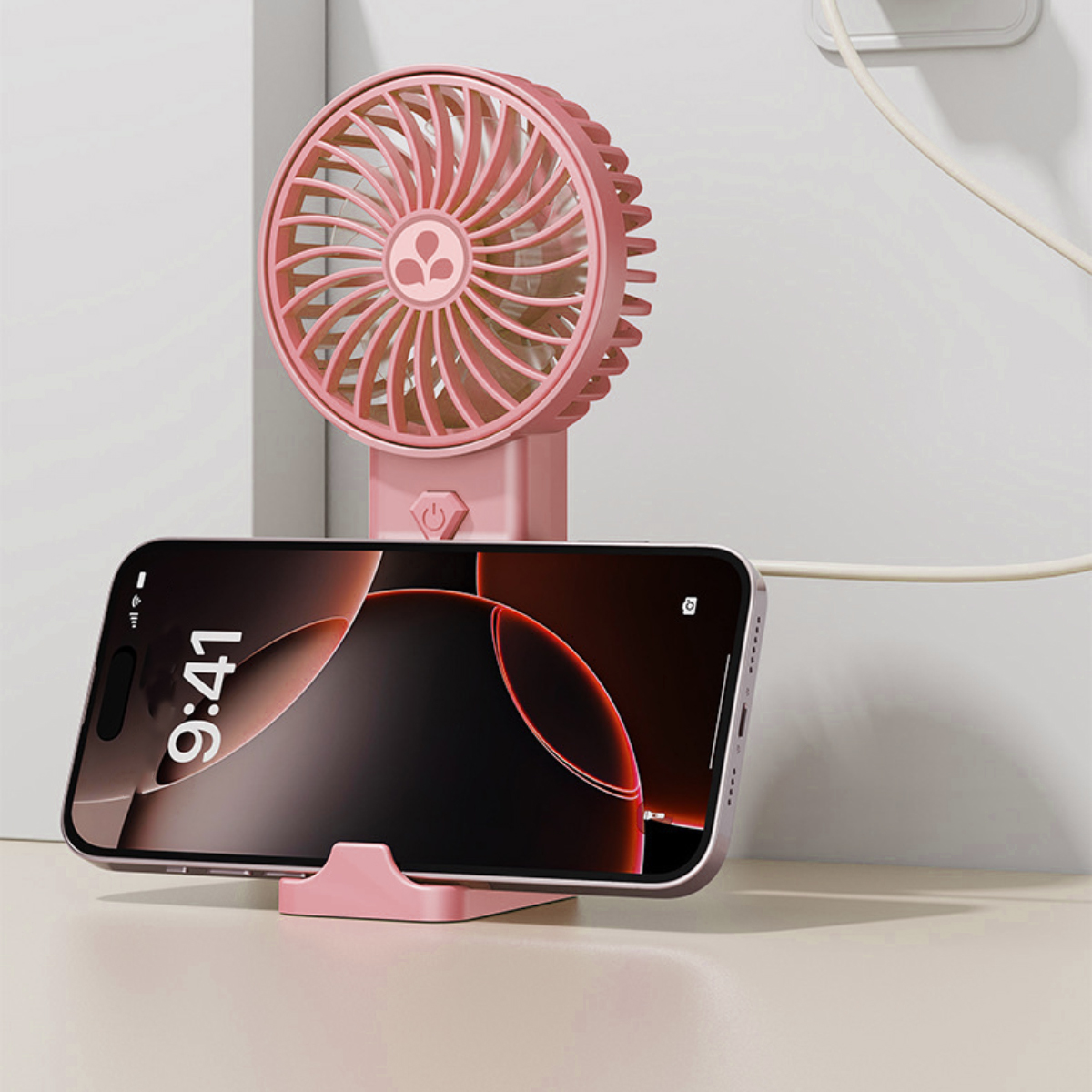 Ein pinker Ventilator steht hinter einem Handy auf einem pinken Ständer. Ein Kabel ist eingesteckt.