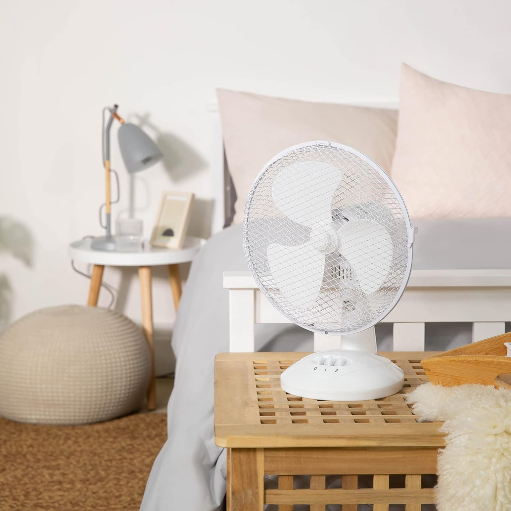 Ein weißer Tischventilator steht auf einem Holztisch neben einem Bett.