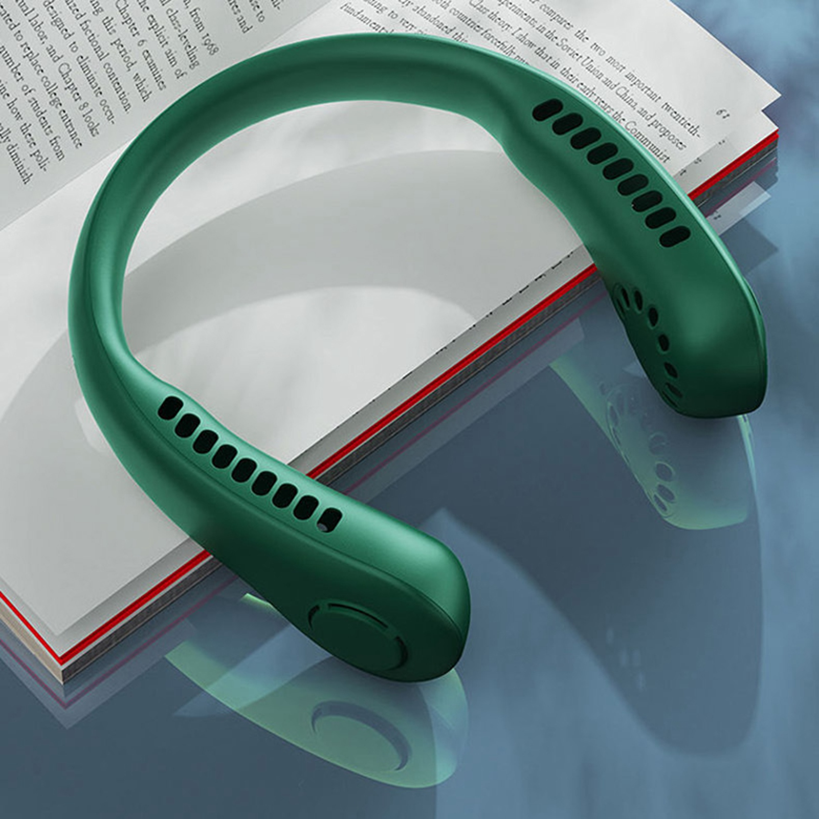 Ein grüner Nackenventilator liegt auf einem Buch auf einem spiegelnden Tisch.